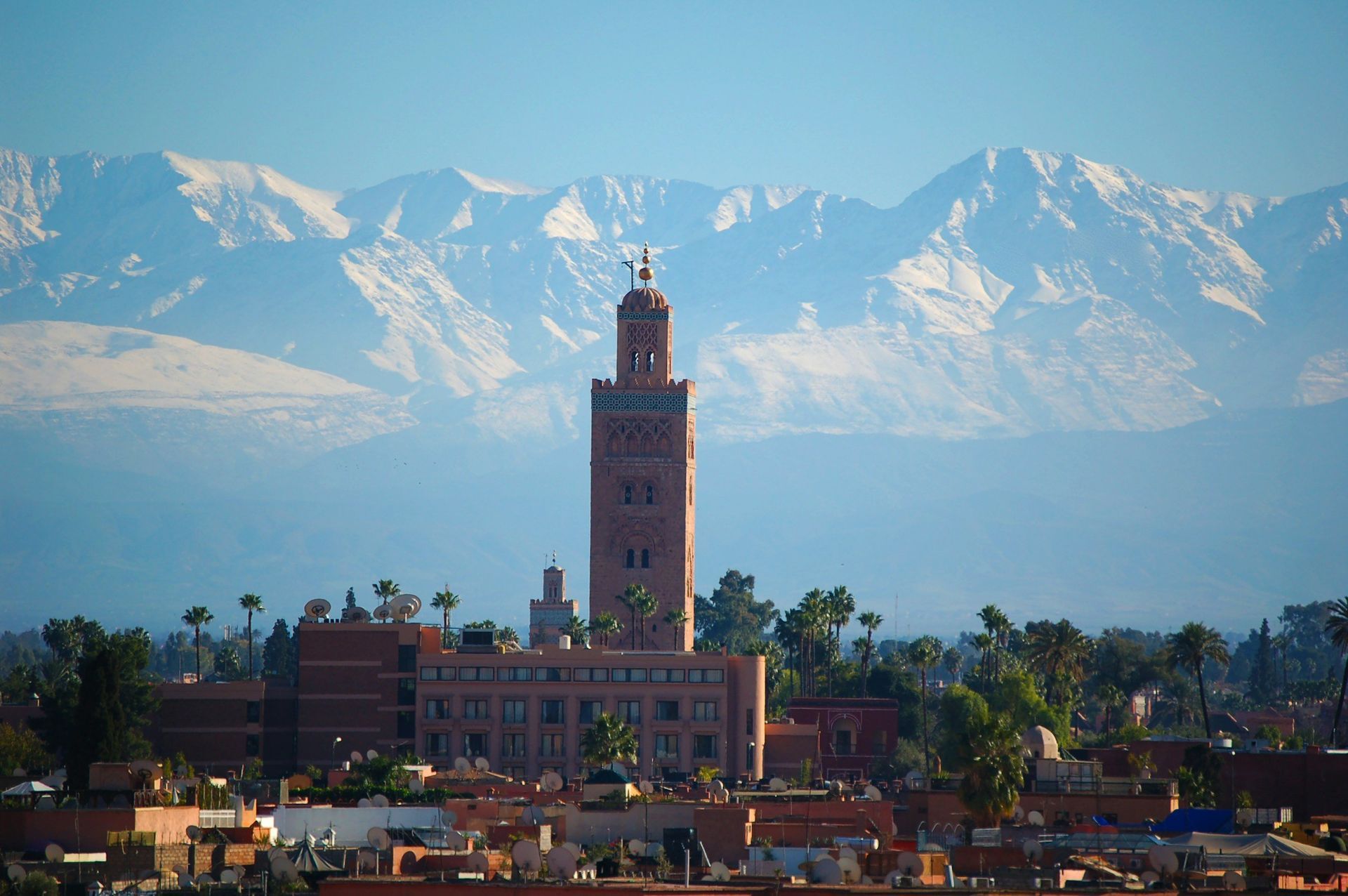 A tall minaret rises above a cityscape, snowy mountains in the background under a blue sky.