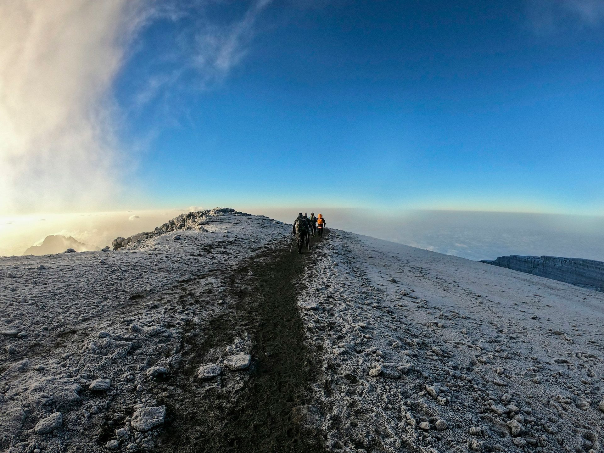 People hike a snowy mountain trail under a bright blue sky, with a view of clouds below.