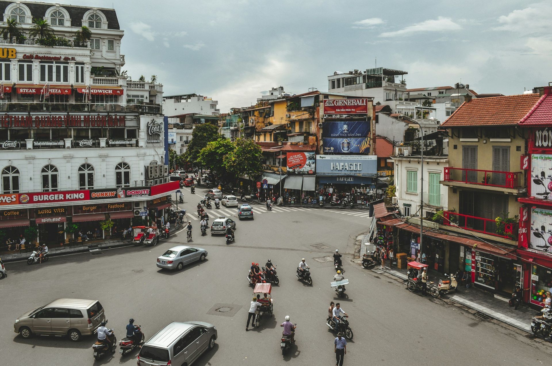 Busy city intersection in Hanoi, Vietnam, with cars, motorbikes, and buildings with red awnings. Overcast sky.