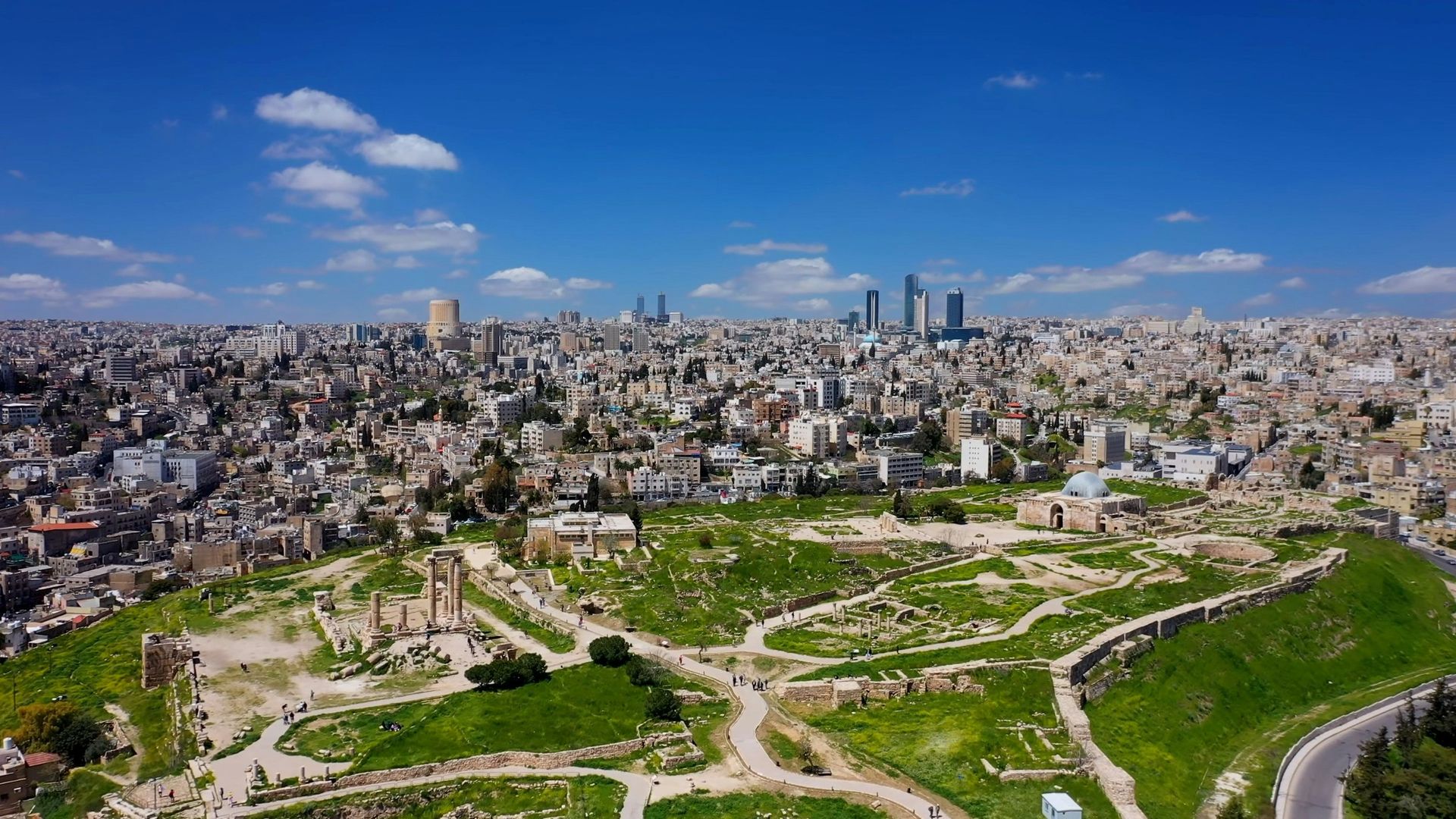 Aerial view of Amman, Jordan, cityscape with ancient ruins in foreground, blue sky and scattered clouds.