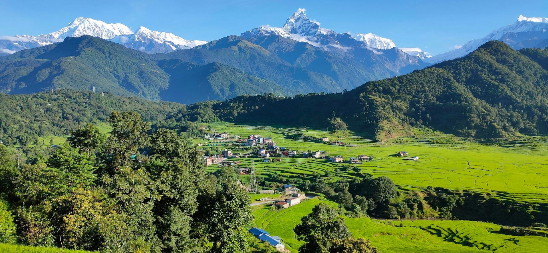 Scenic mountain landscape with snow-capped peaks, green hills, and a village under a clear blue sky.