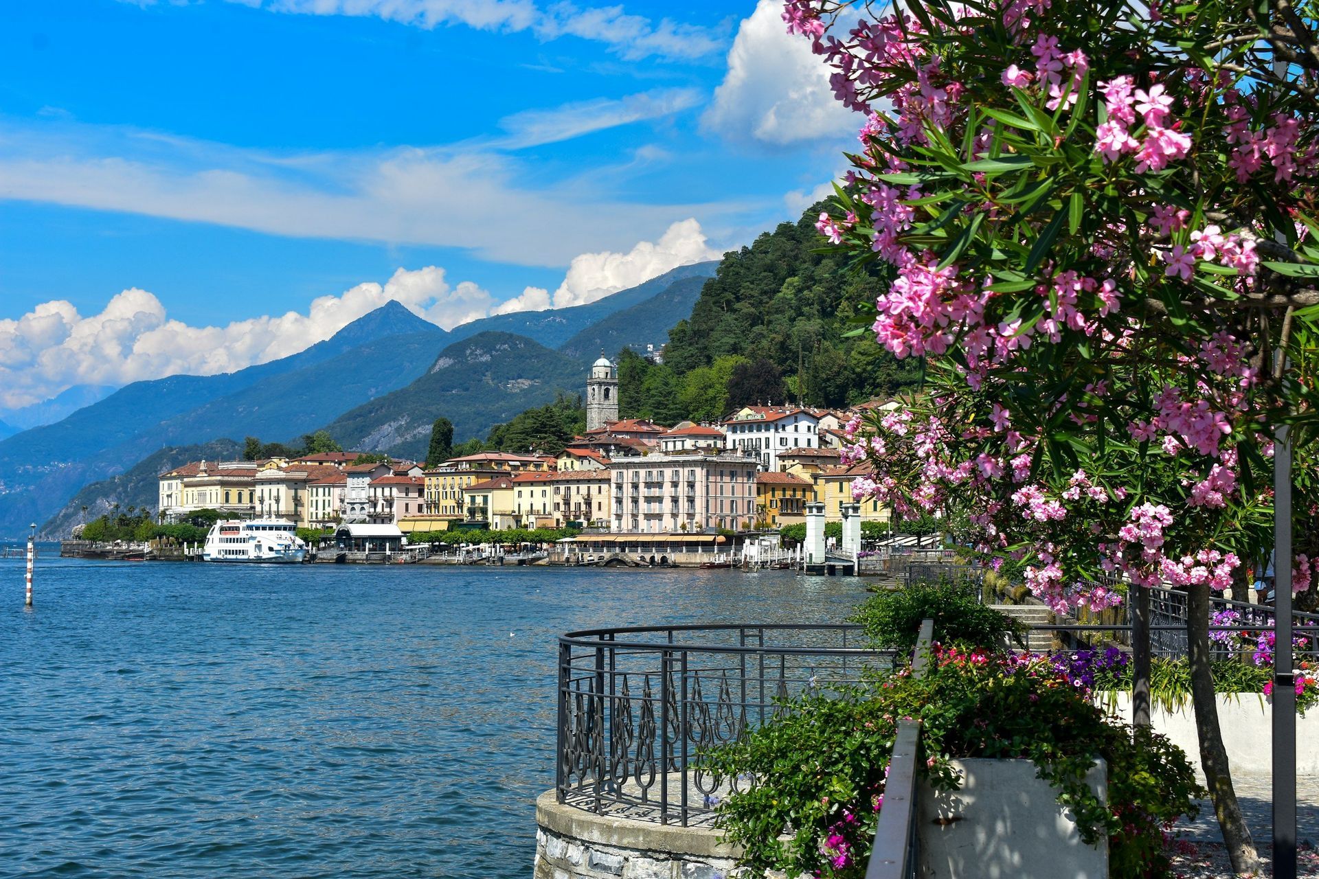Lake Como town with water, buildings, mountains, blue sky, and pink flowers.