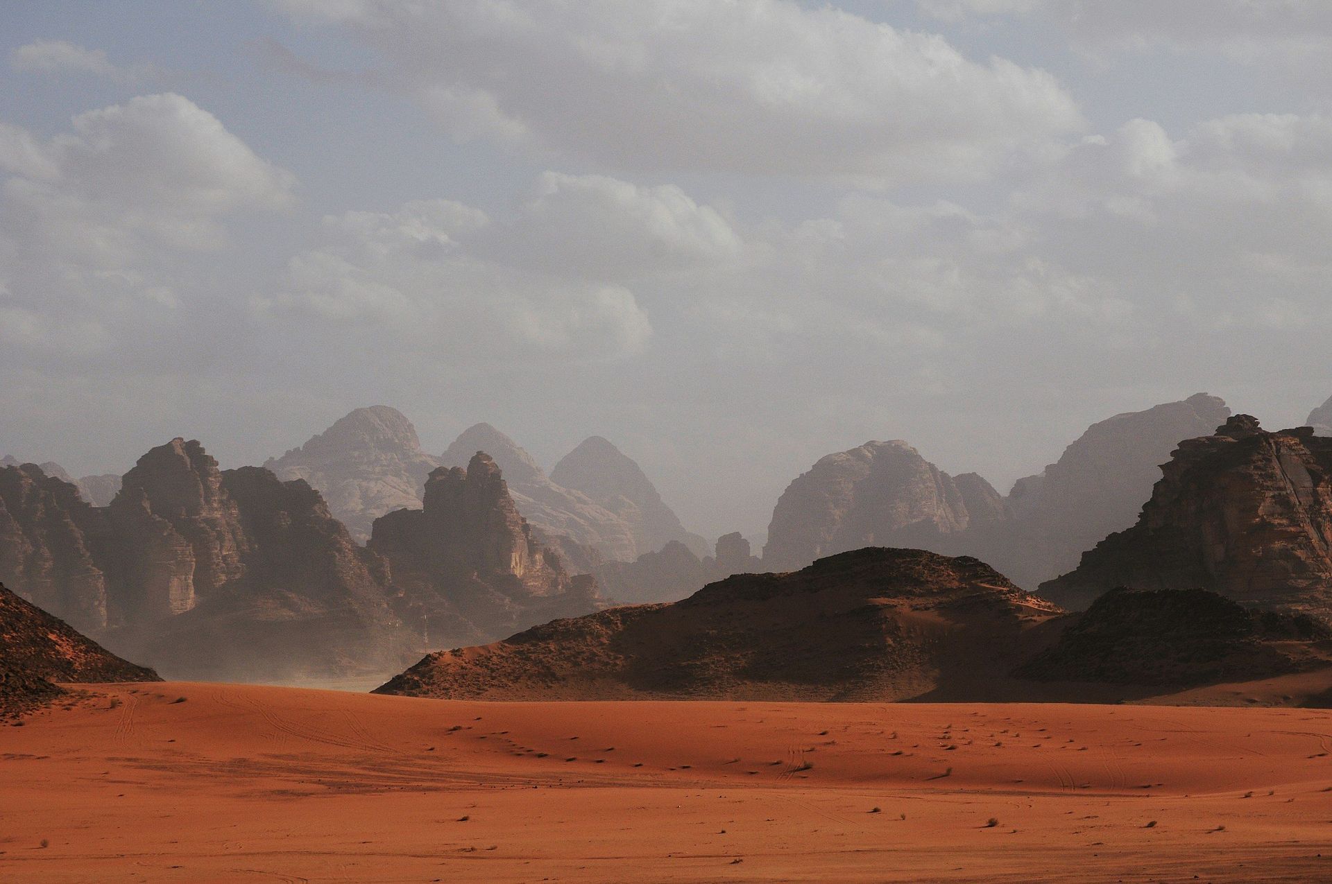 Red desert landscape with distant rocky mountains and a cloudy sky.