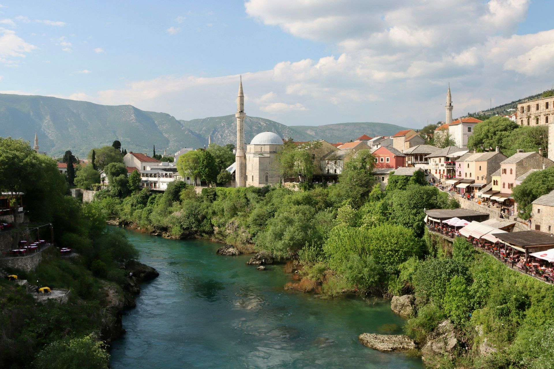 Mostar, Bosnia-Herzegovina: A river flows through a historic town, with a mosque and buildings set against a mountain backdrop.