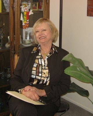 Smiling woman seated indoors in a black blazer, holding papers beside a glass cabinet and large green plant