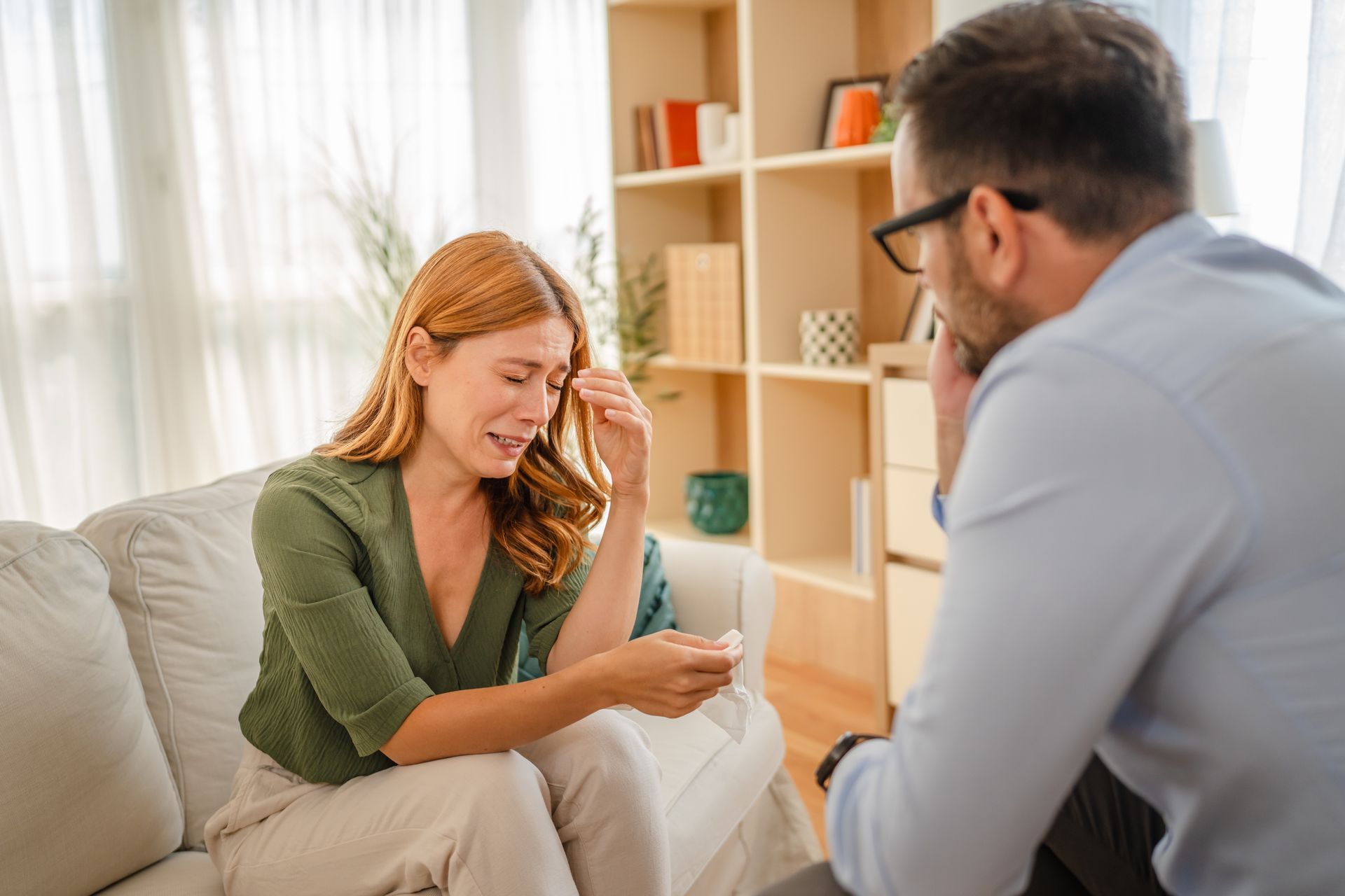 Two people talking on a couch in a bright living room, one holding a smartphone.