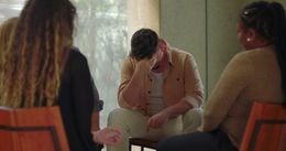 Three people sit in a circle, one looking down with head in hands, in a softly lit room.