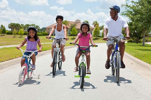 Family of four riding bicycles on a paved road, wearing helmets, sunny day.
