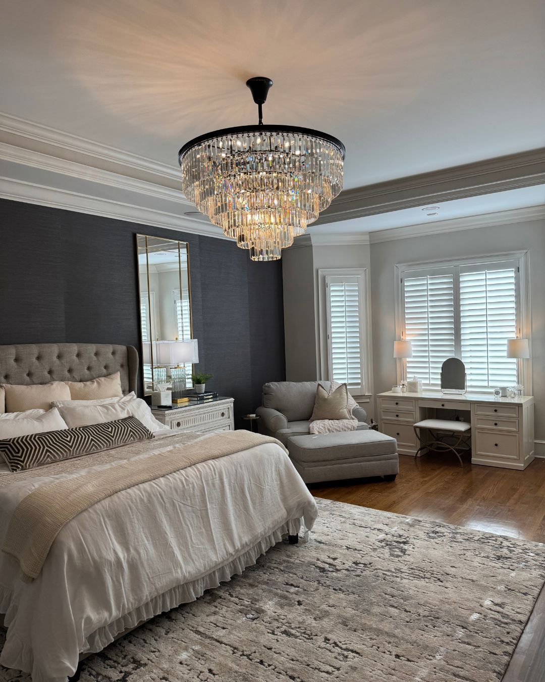 Elegant bedroom with crystal chandelier, tufted bed, chaise lounge, and white desk by window shutters
