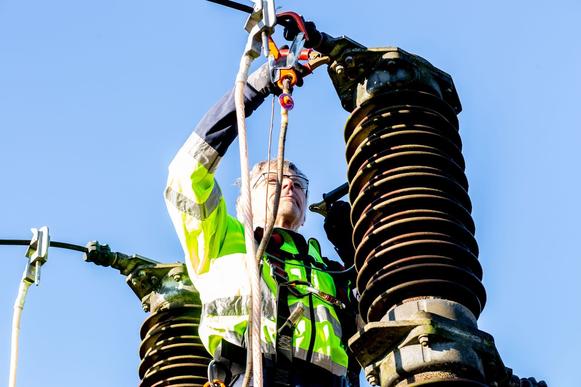 Worker in a neon safety jacket repairing electrical equipment on a utility pole under a blue sky