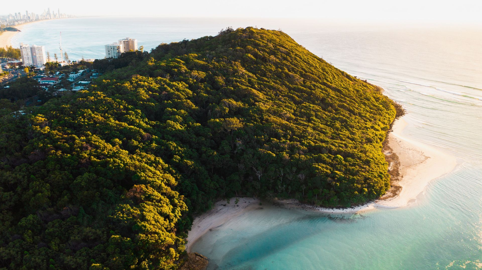 A lush green tropical peninsula curves into turquoise water beside a sandy beach at sunrise.