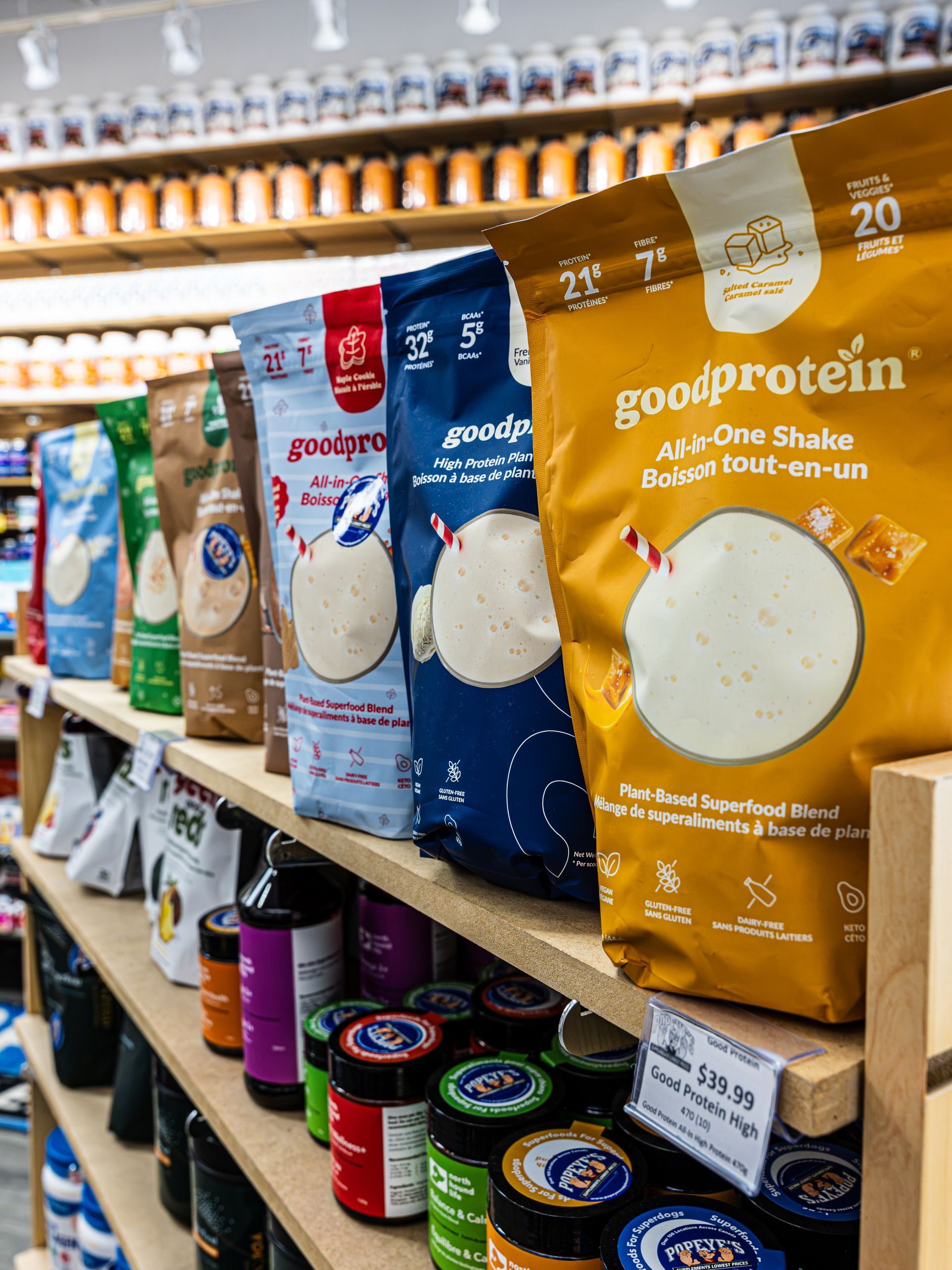 Grocery store shelf with colorful protein powder bags and tubs, including Good Protein almond vanilla shake mix