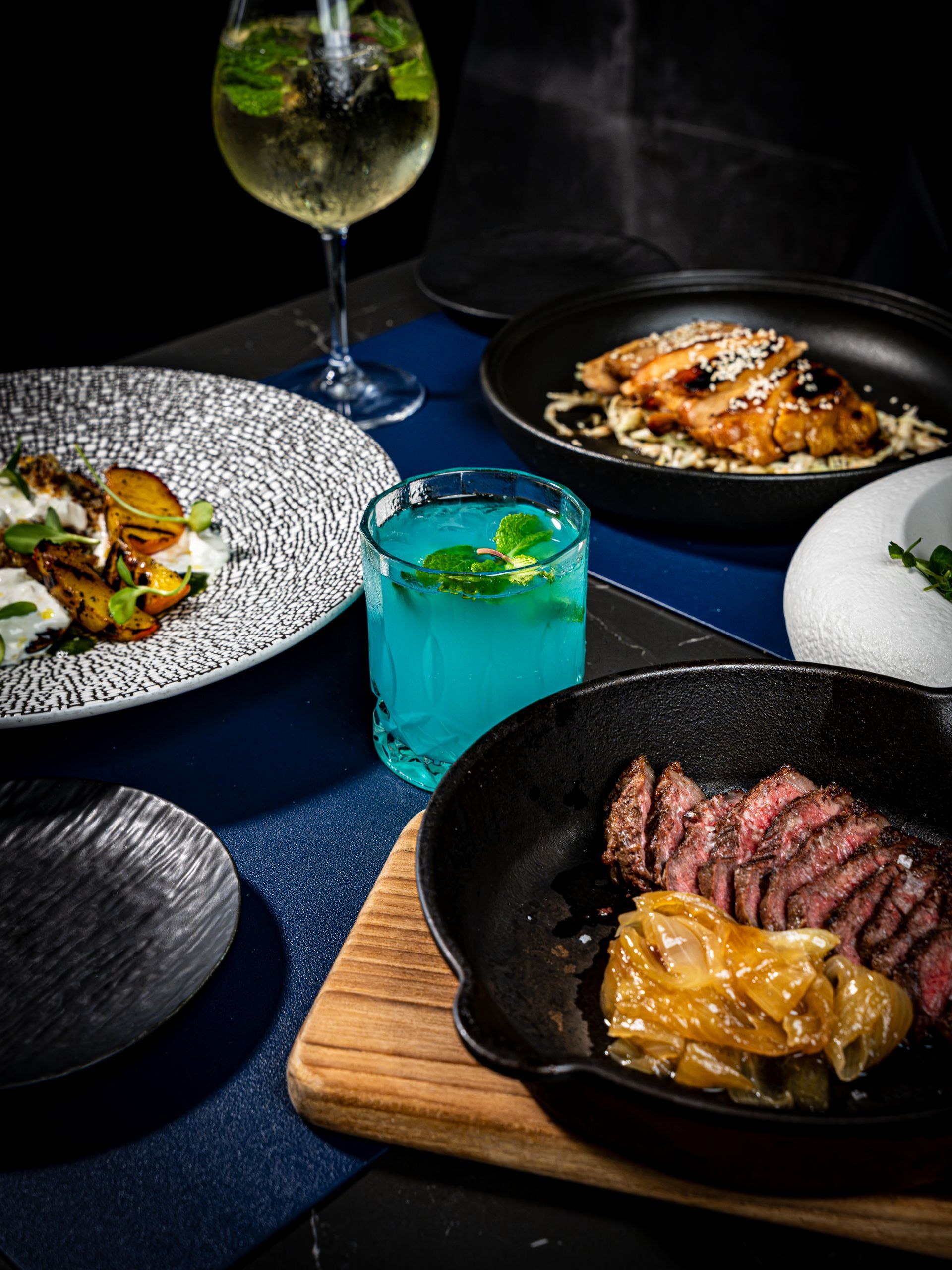 Plated gourmet dishes and cocktails on a dark blue table in a dimly lit restaurant setting