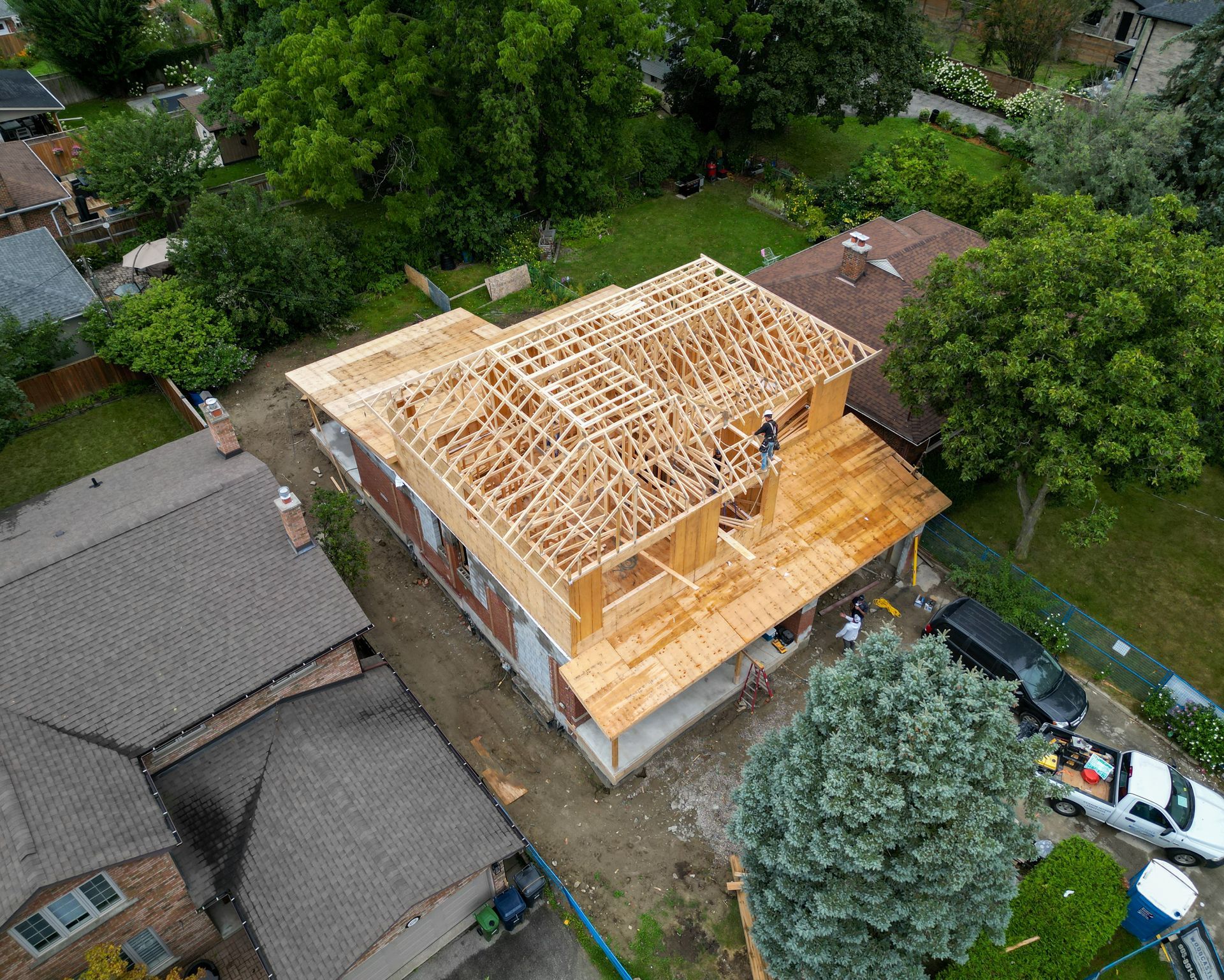 Aerial view of a house under construction with a new wooden roof frame, surrounded by trees and neighboring homes