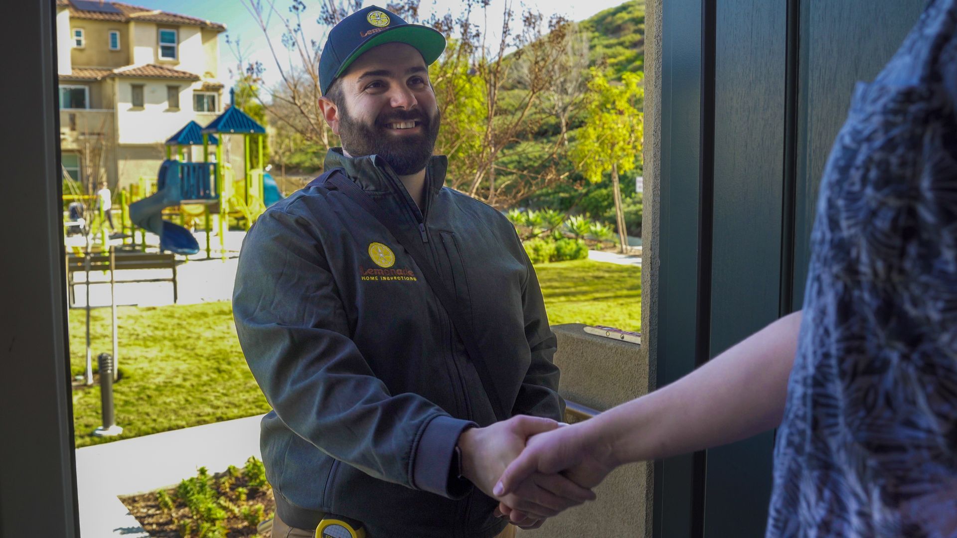 Person in uniform shakes hands at a doorway, smiling. Playground and houses visible in the background.