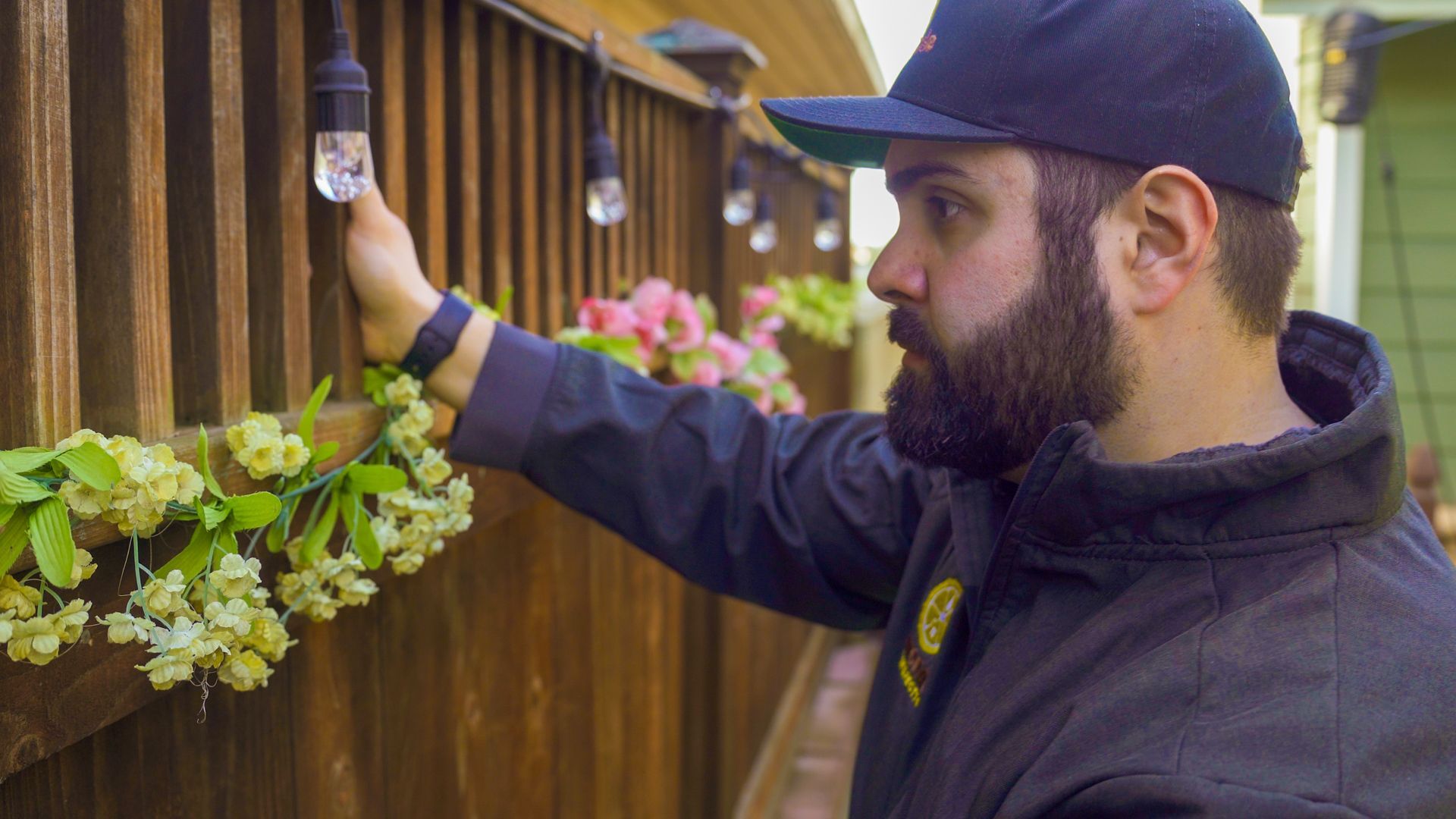 Man in a black cap and jacket painting a wooden fence beside hanging flowers