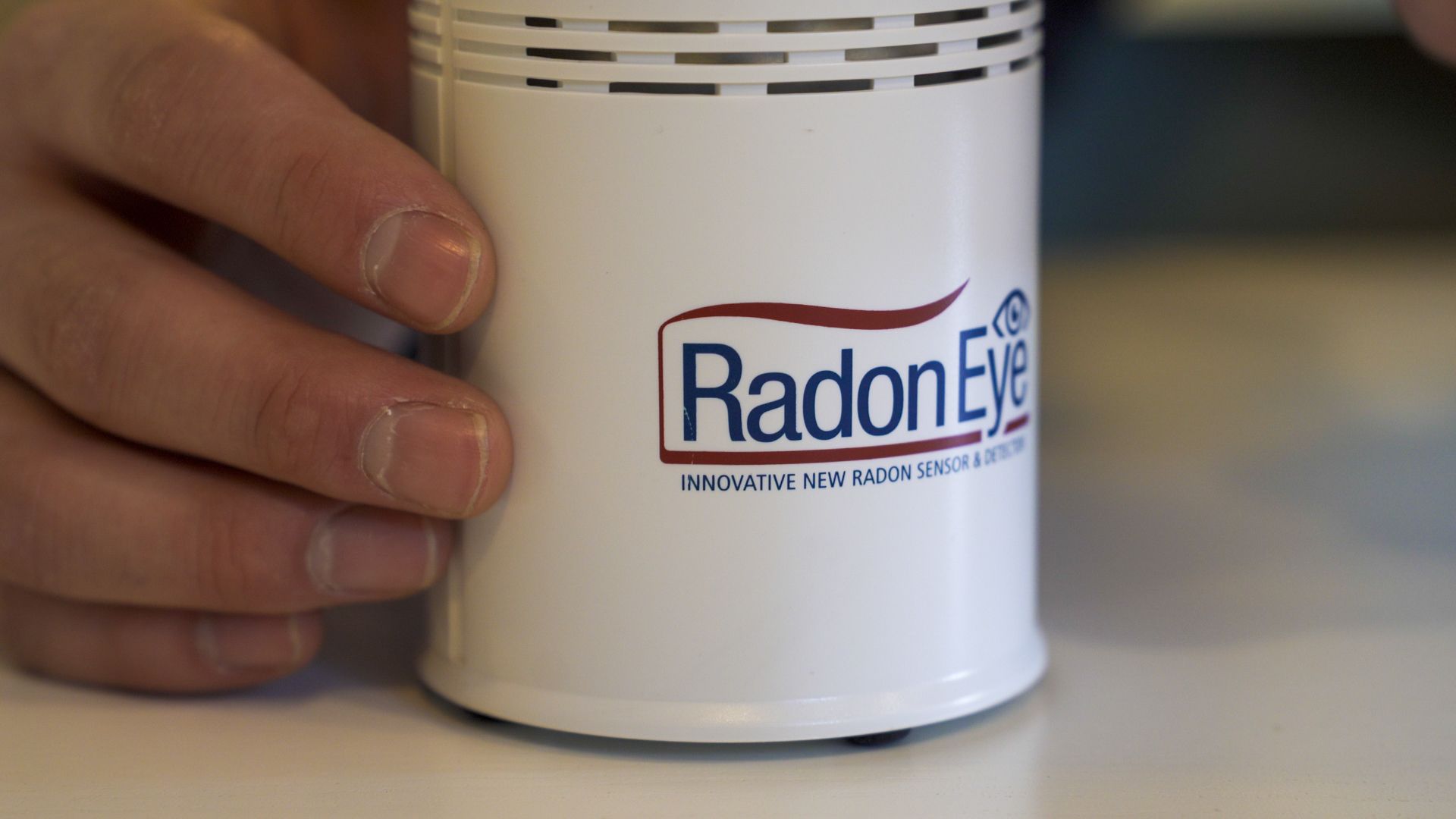 Hand holding a white RadonEye device with a logo on the front