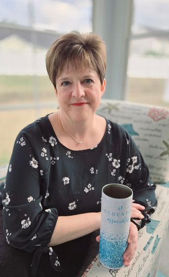 A person with short hair sits in a chair, wearing a black floral top, holding a blue and white tumbler.