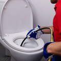 A plumber in a red uniform and work gloves uses a drain snake to unclog a white toilet.