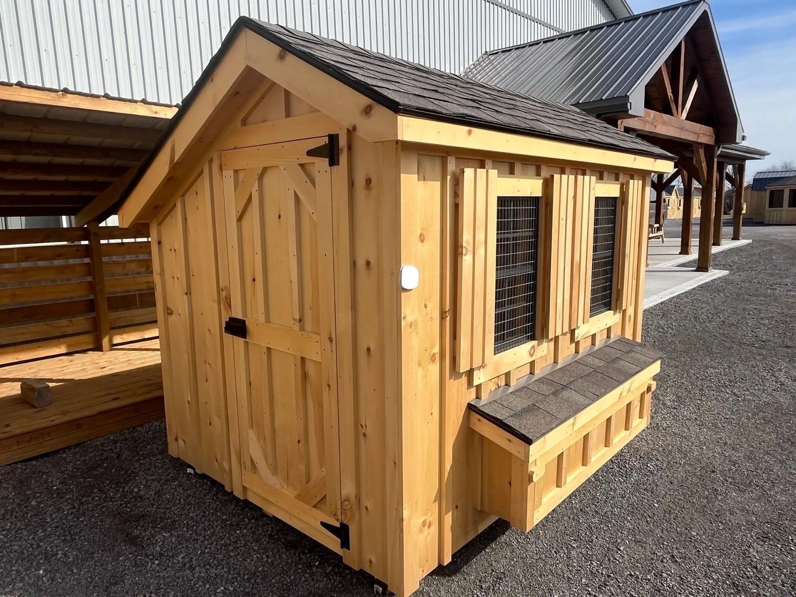 Small wooden chicken coop with wire window and slanted roof on gravel beside a larger shed
