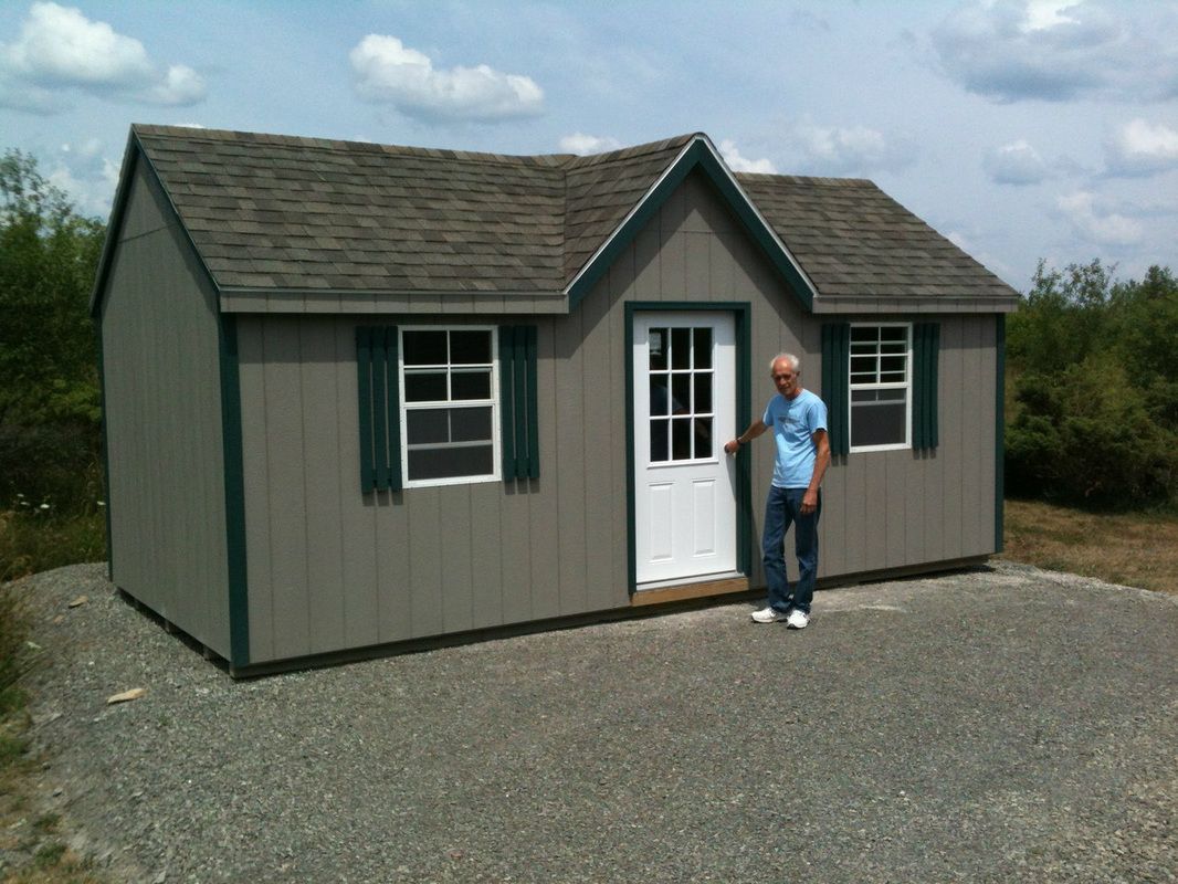 Small gray shed with white door and windows on gravel, with a person standing beside the entrance.