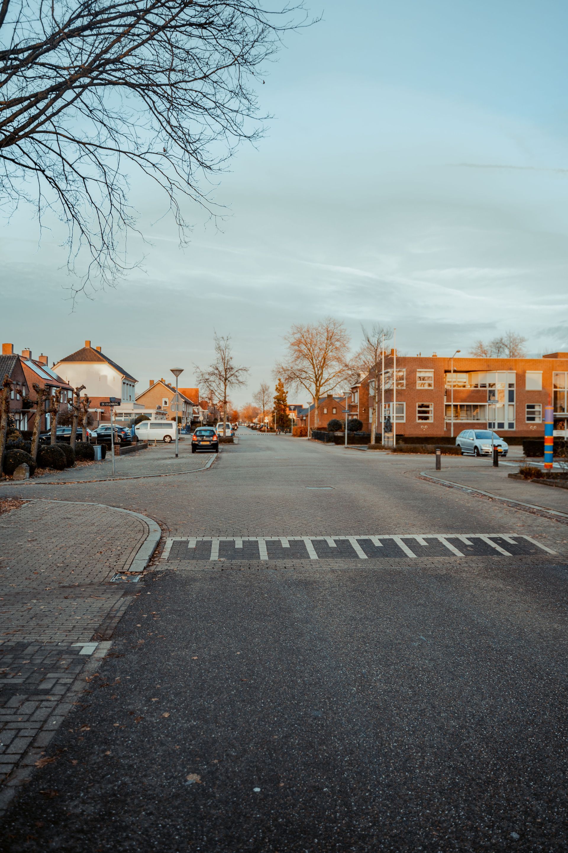 Quiet residential street at dusk with parked cars, brick buildings, and bare trees under a pale sky