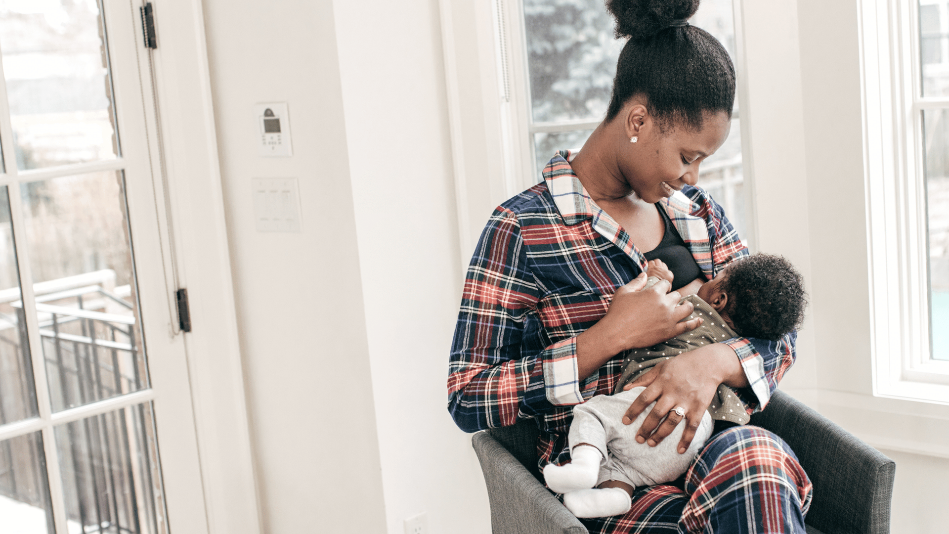 Mother breastfeeds her baby while seated in a chair by a bright window.