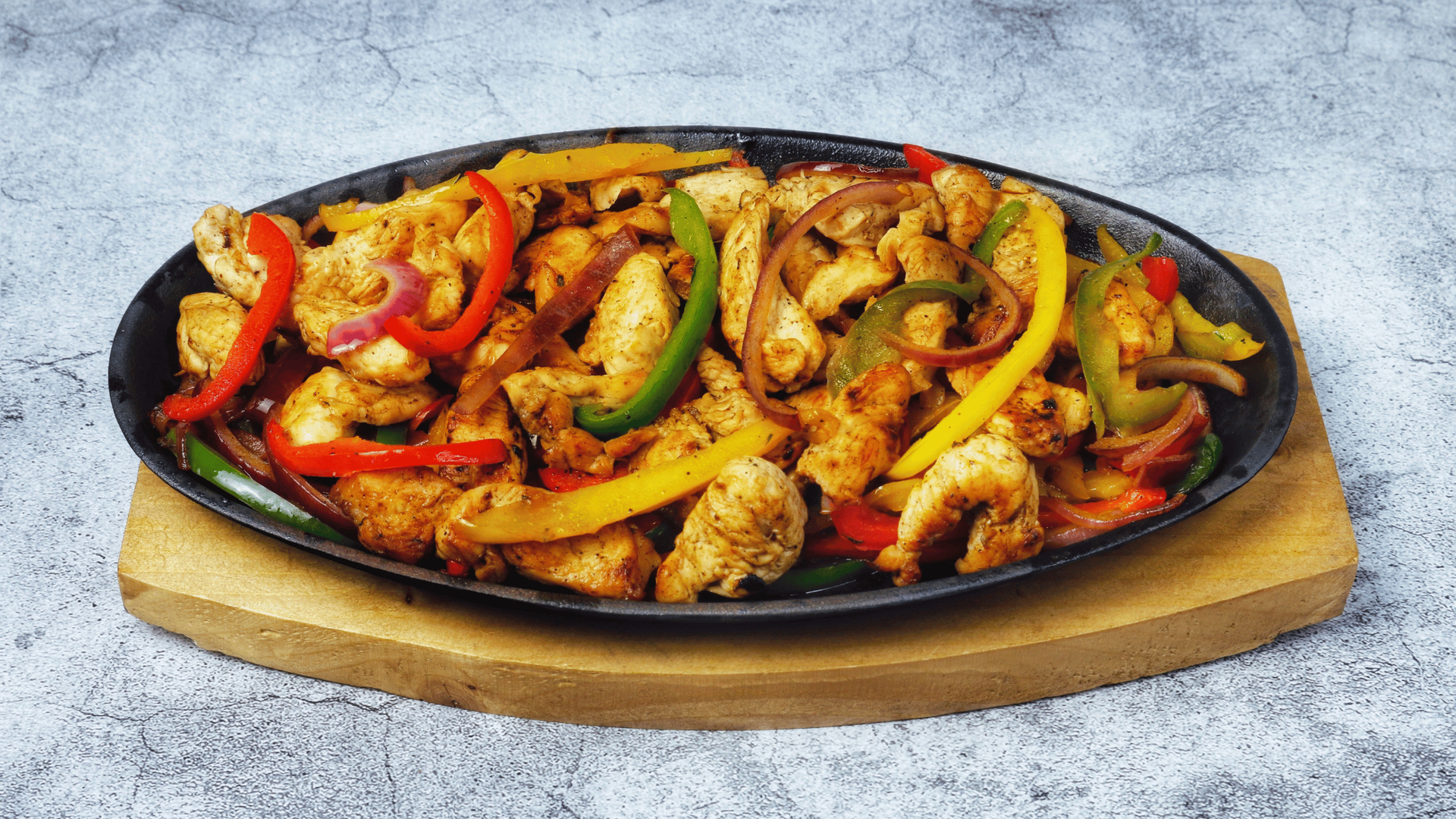 Chicken fajitas in a black skillet, placed on a wooden board, with vibrant peppers and onions.