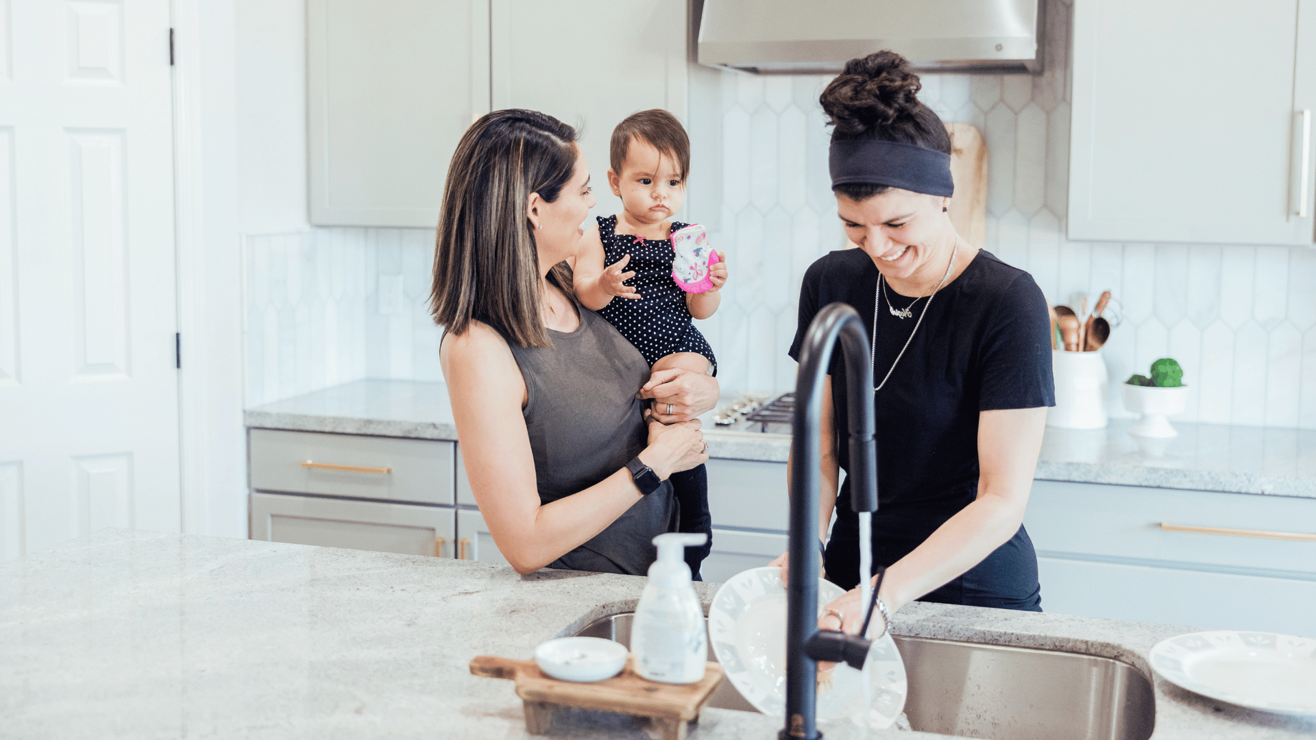 A woman holds a toddler in her arms in a bright kitchen, smiling as another woman washes dishes.