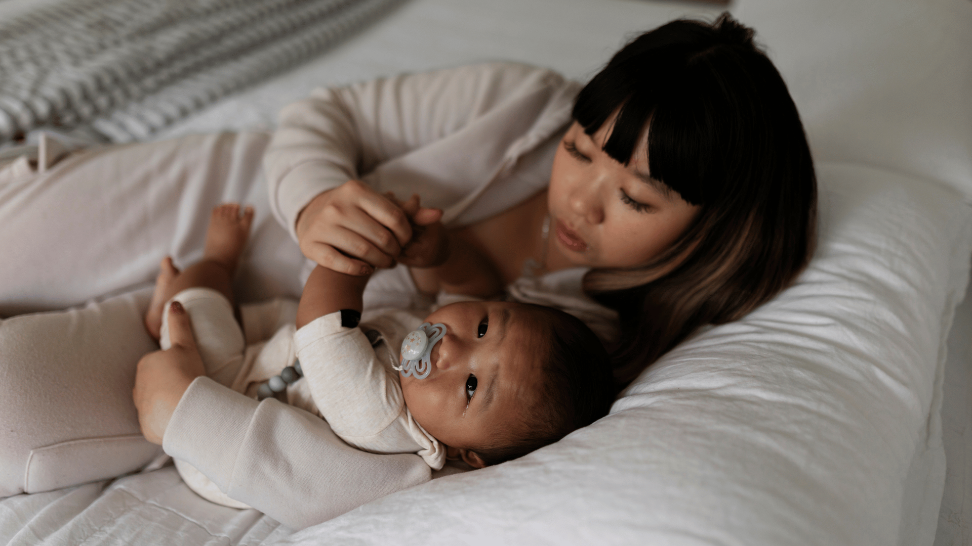 A mother and baby on a bed, with the mother holding the baby's hands and looking down.