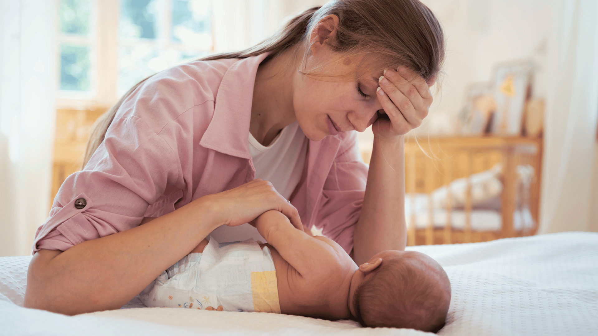 A tired mother in a pink shirt rests her head on her hand while tending to her baby on a bed.