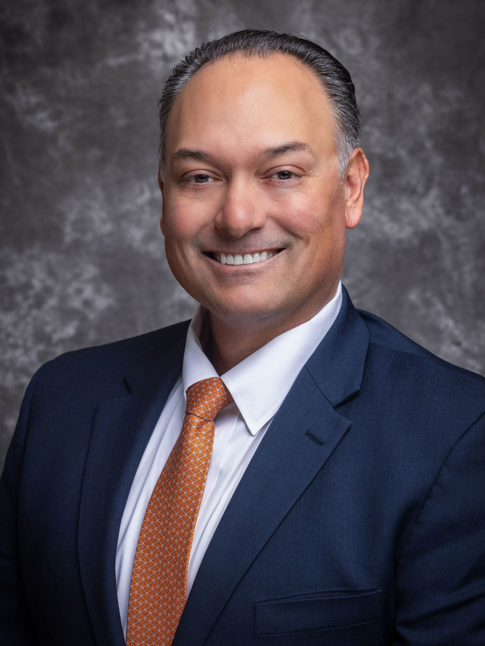Man in blue suit, orange tie, smiling. Studio background.