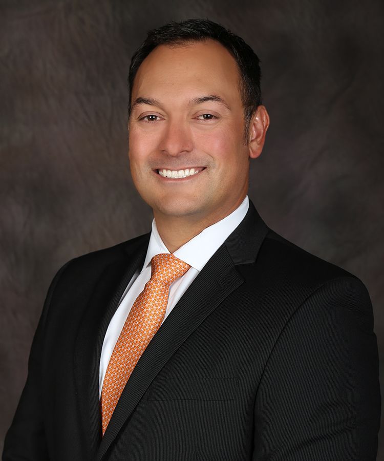 Man in black suit and orange tie smiling, against a dark background.