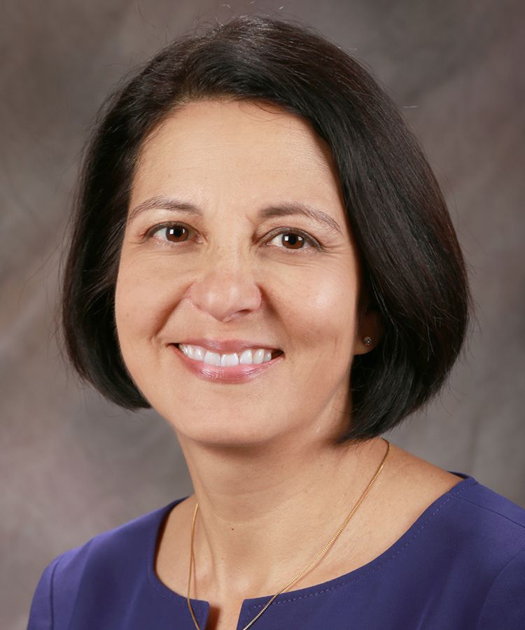 Woman with dark hair smiles, wearing a purple top, against a neutral background.