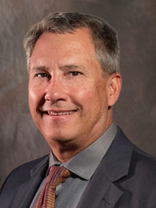 Man in a suit smiles, gray hair, and a patterned tie, against a dark background.