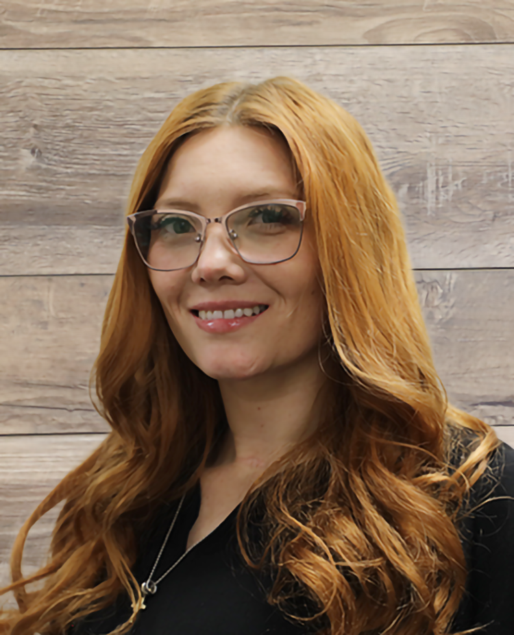 A woman with red hair and glasses is smiling in front of a wooden wall.