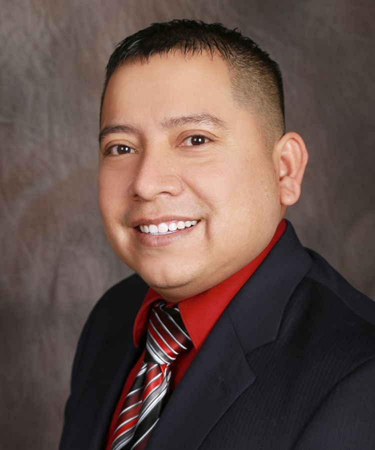Man in suit smiles at the camera, red shirt and patterned tie. Against a dark, blurred backdrop.