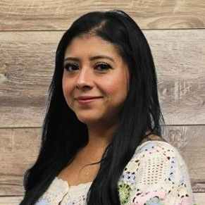 Woman with dark hair, smiling, wearing a patterned sweater, against a wood-paneled wall.