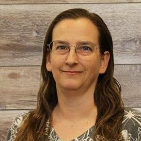 Woman with glasses smiles; light brown hair, patterned shirt, wooden wall background.