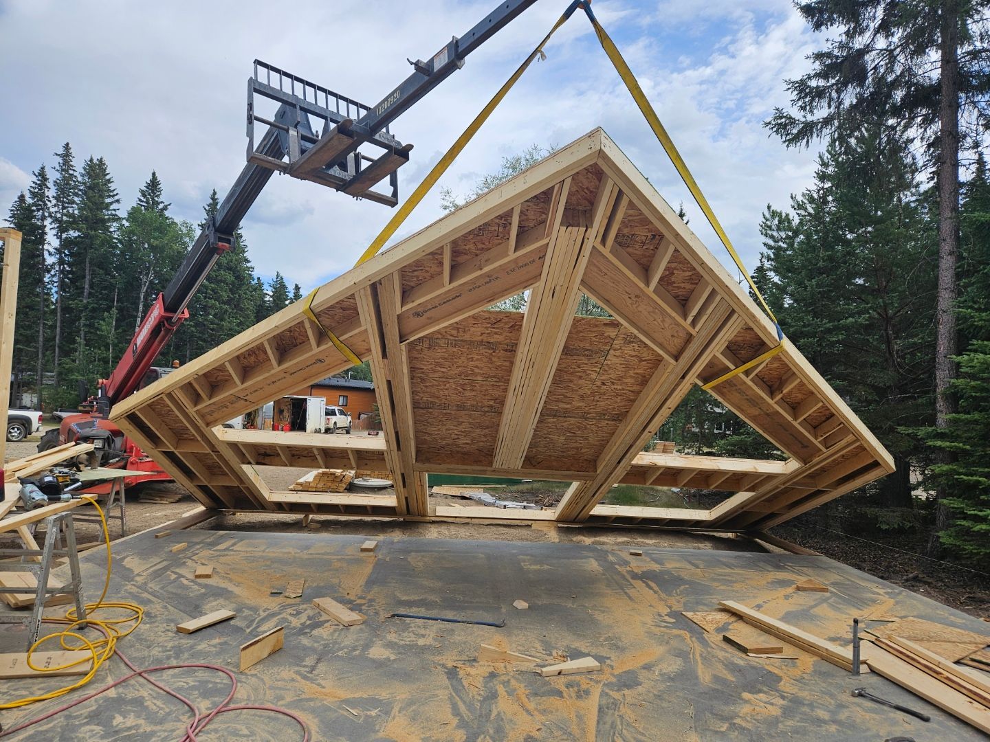 A framed roof section being lifted by a telehandler crane at a construction site.