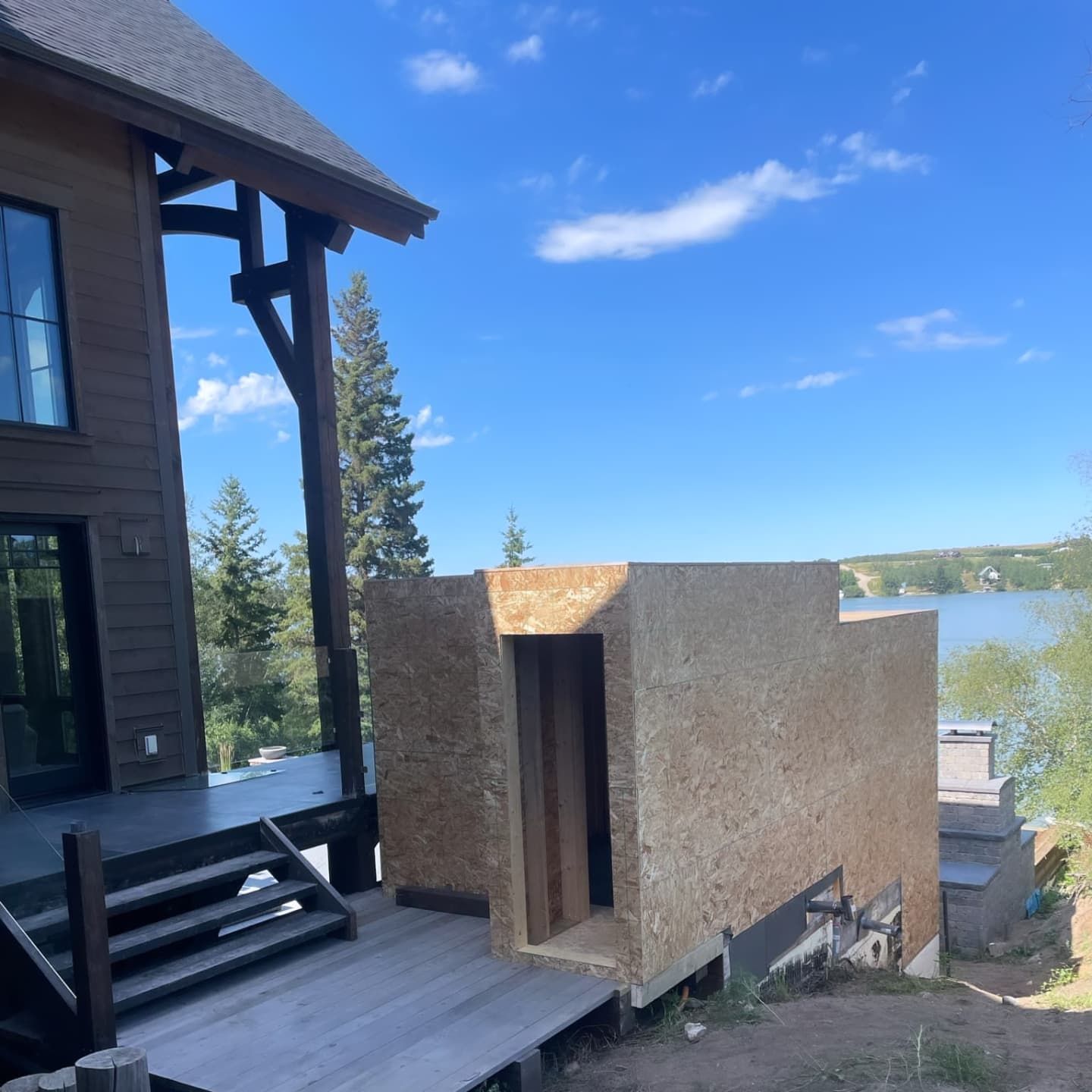 A wooden structure is being built next to a house with a deck overlooking a lake and blue sky.