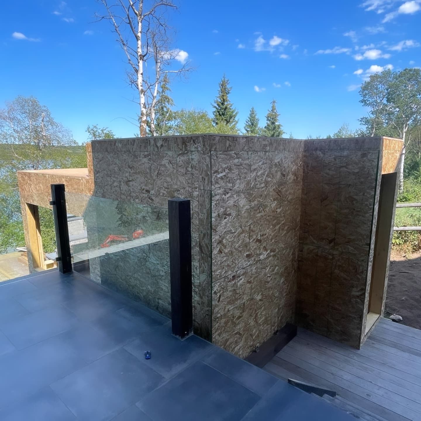 Exterior view of a deck under construction with OSB walls, glass panels, and a blue sky in the background.