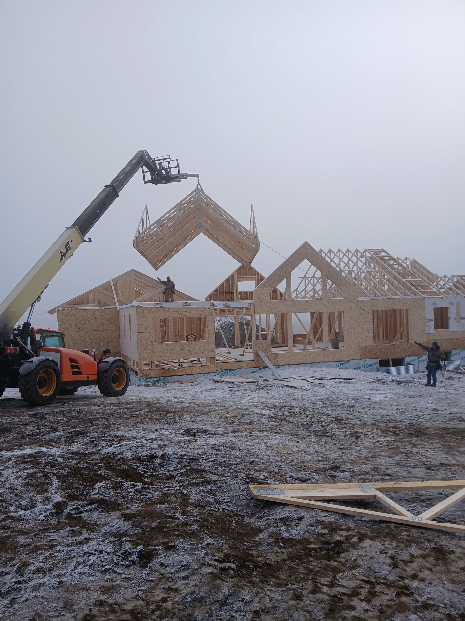 Construction site: A telehandler lifts a roof truss onto a wood-framed house under a foggy, snowy sky.