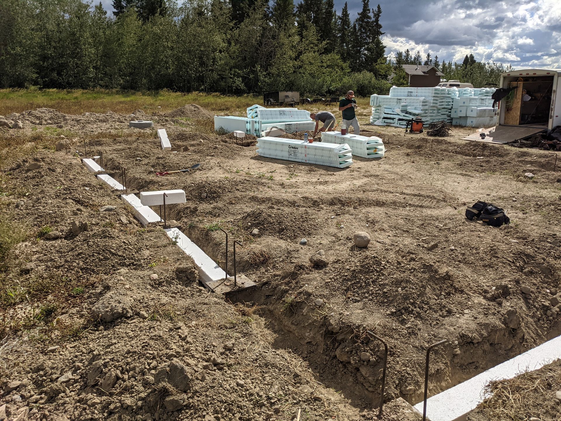 Construction site with workers, foam insulation, and foundation trenches. Outdoors, daylight.