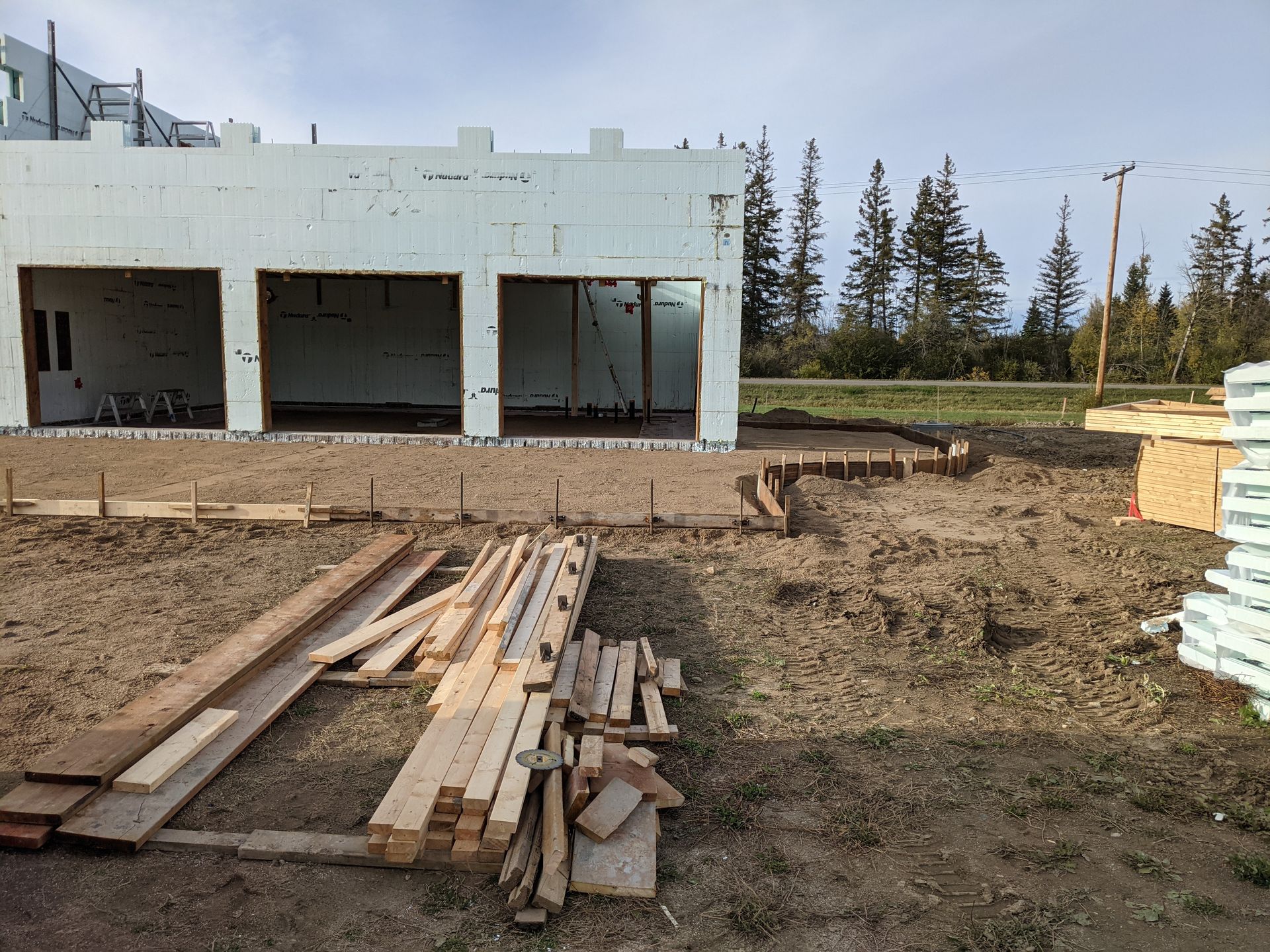 Construction site: Foam block walls, wooden framing, lumber pile, dirt ground, and trees in the background.