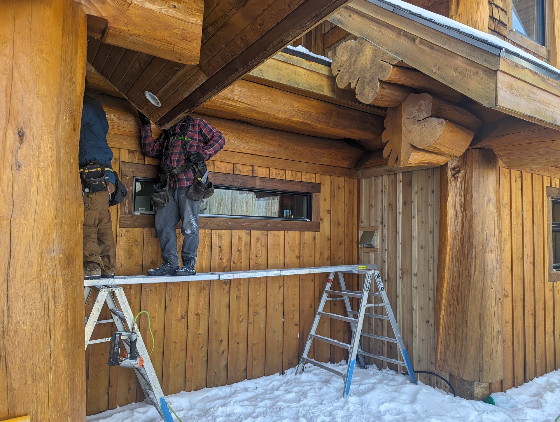 Two people on a log cabin roof, one on a ladder, working on the exterior in the snow.