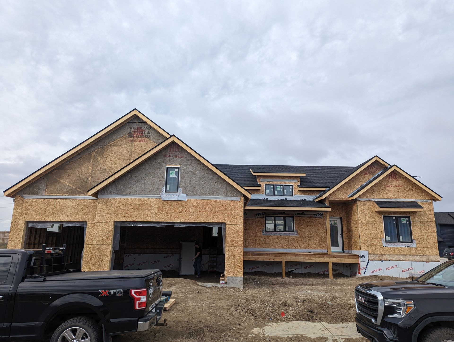 House under construction, with exposed wood framing, garage, and two trucks parked out front under a cloudy sky.
