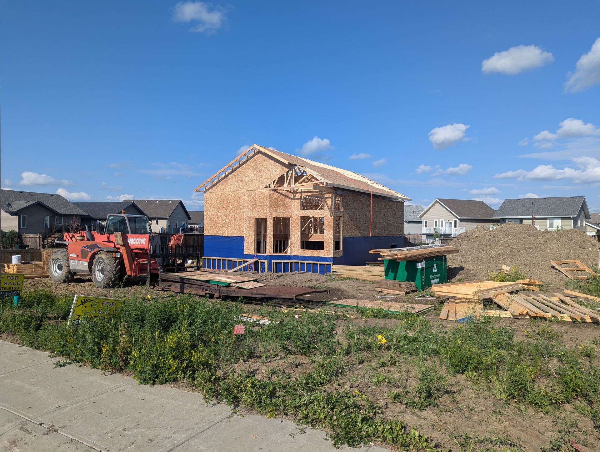 Construction of a new house; wooden frame, blue wrap, and red tractor in a residential area on a sunny day.