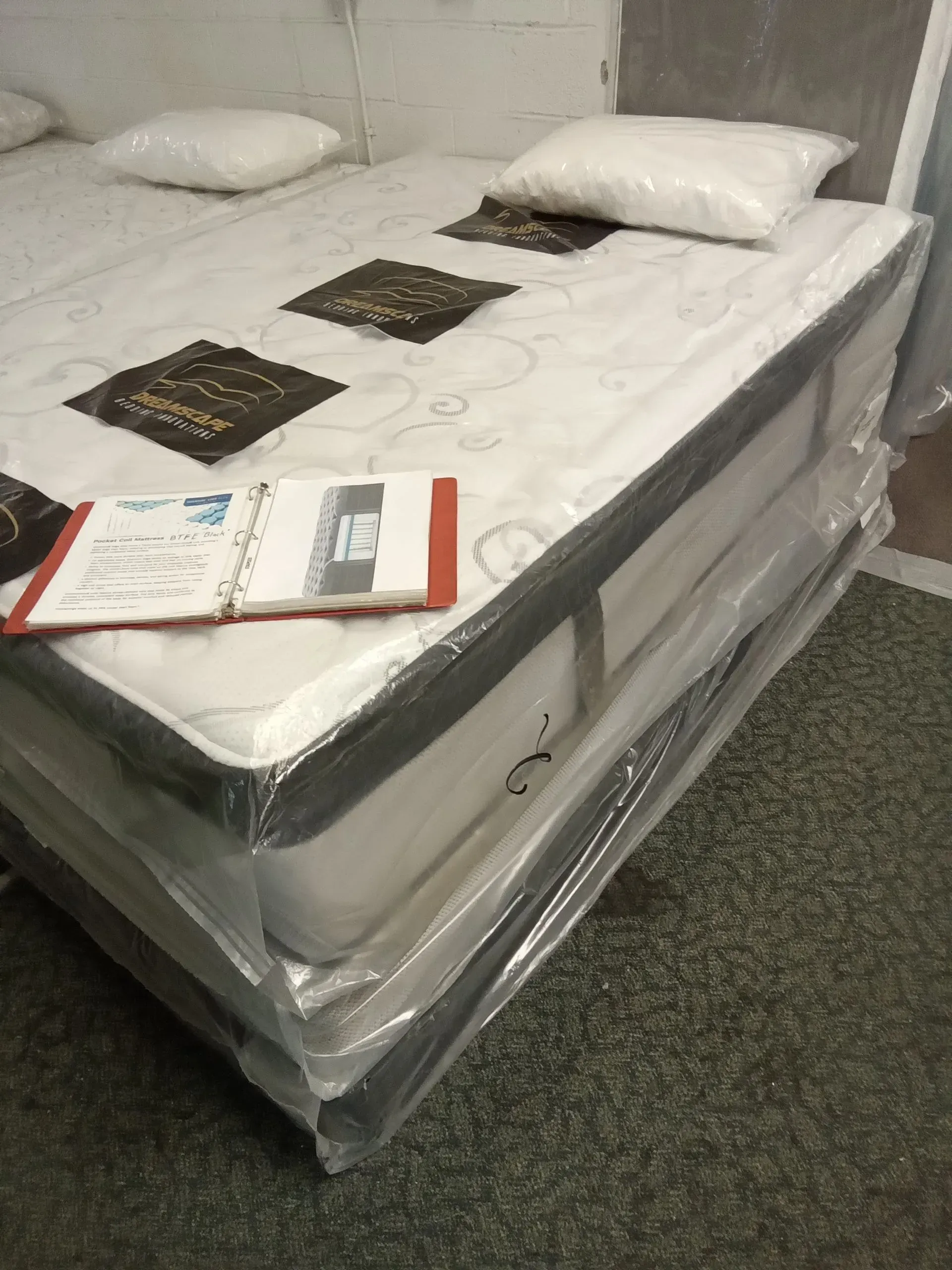 Wrapped mattress on a bed frame with pillows and brochures in a store or warehouse setting
