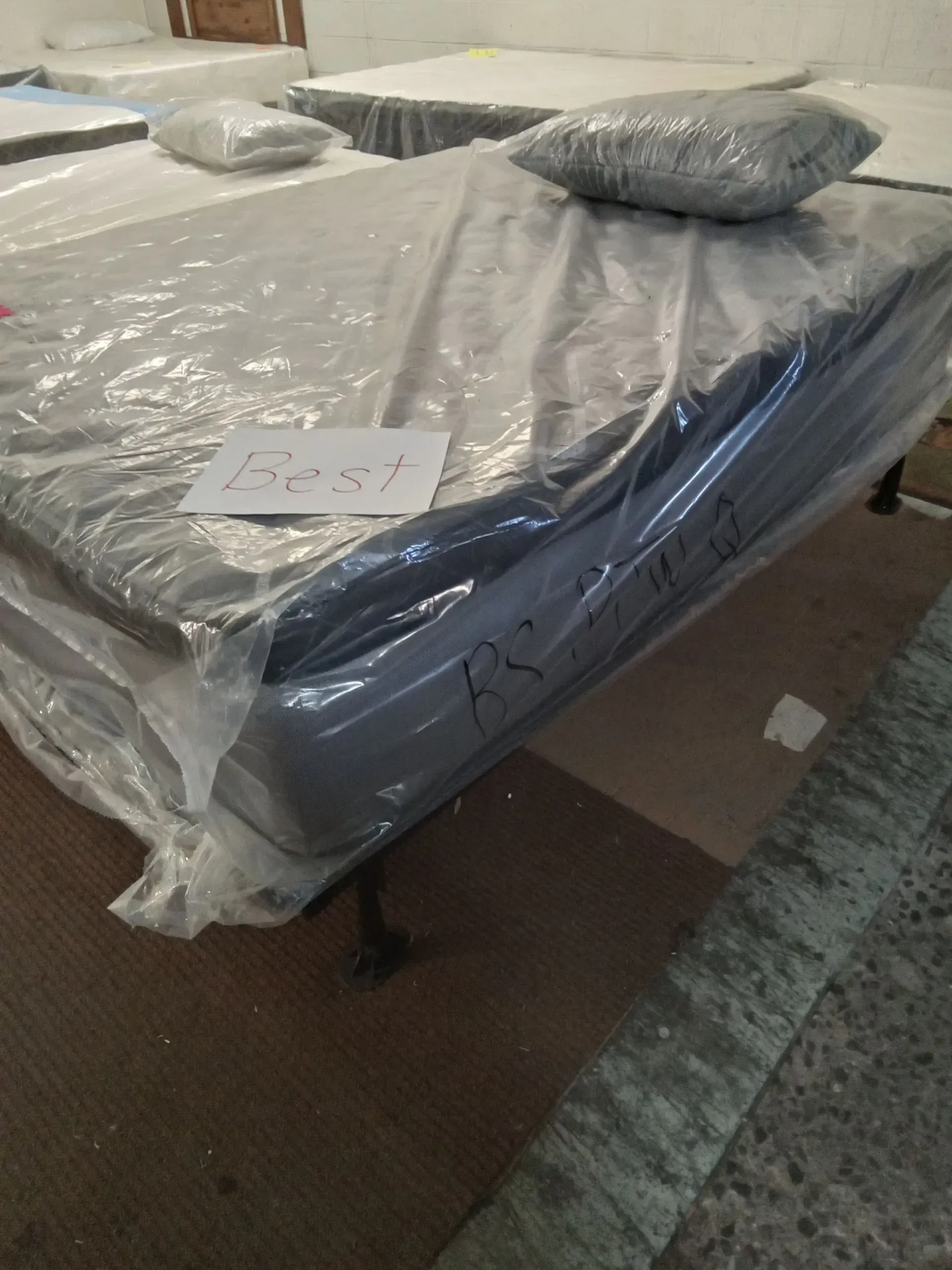 Plastic-wrapped gray bed mattress with a pillow on top, in a room with brown carpet.